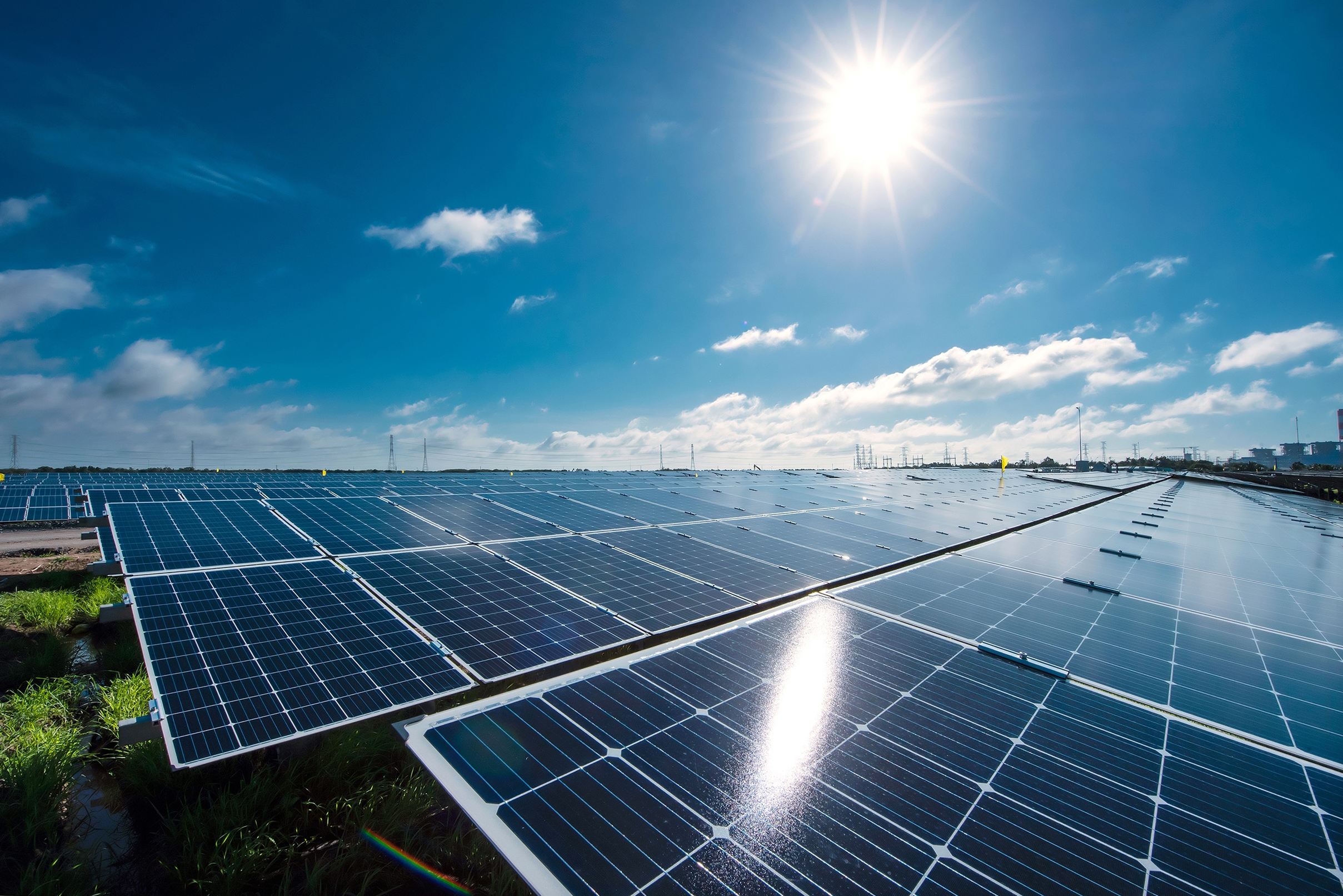 Solar panes in a solar park used for clean energy production. Solar panel under blue sky with sun. Green grass and cloudy sky. Alternative energy concept to reduce global warming and climate change